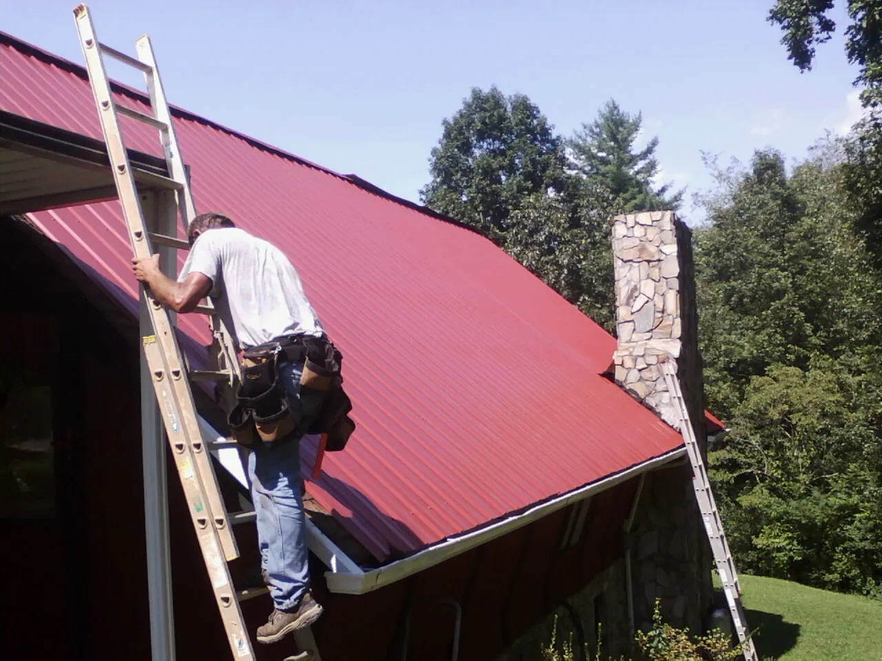 Roofer on a ladder beside a red metal roof and stone chimney — Soffit & Fascia Repair by Roofing Brothers in Bettendorf
